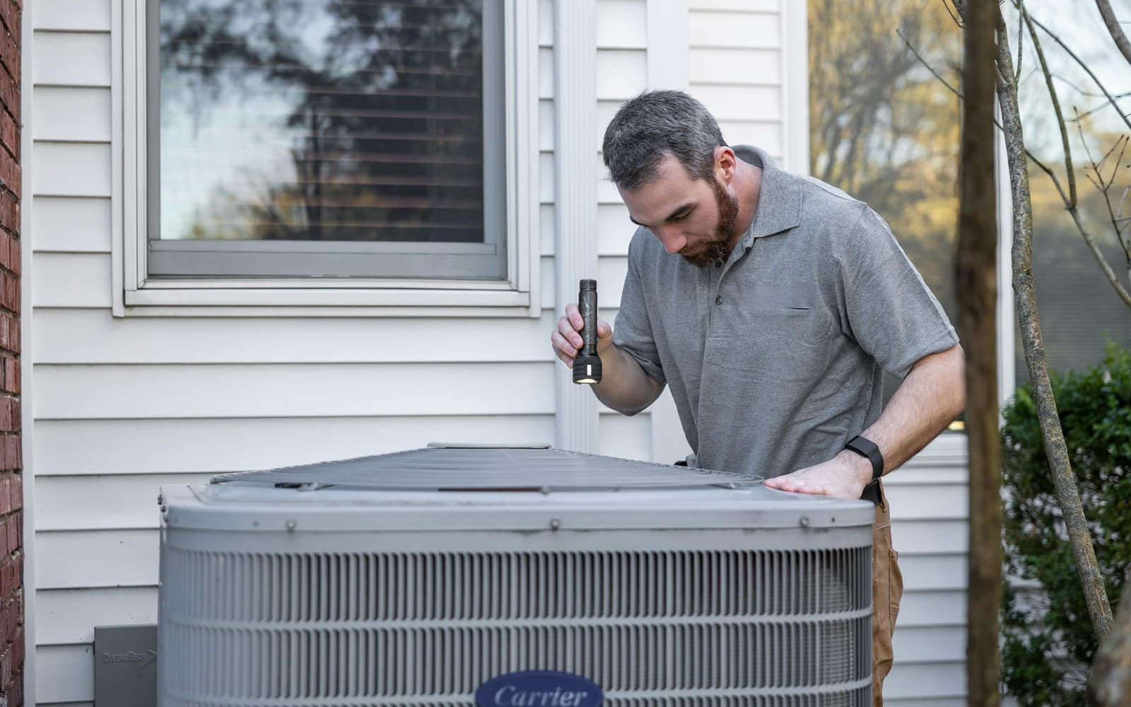 Aces Highlands technician inspecting an outdoor heat pump condenser unit beside a Southern Maine home