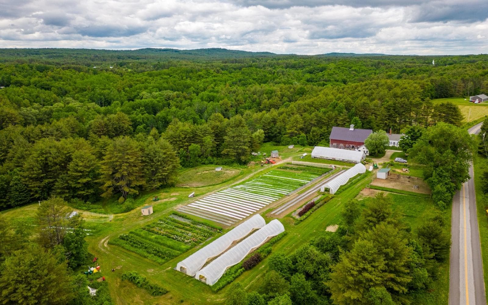 Aerial view of Eastern River Farm's barn, greenhouses, and flower rows surrounded by forest in Pittston, Maine